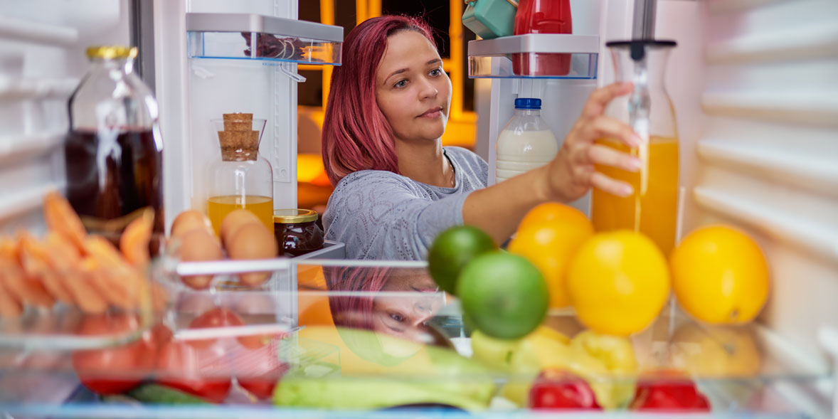 Woman with pink hair reaching for a bottle of juice inside an open refrigerator filled with fruits and other beverages.