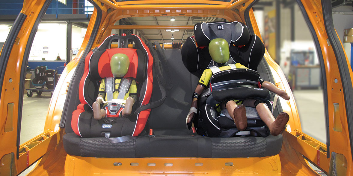 Two child safety seats in the back of an orange van, each holding a crash test dummy with green heads.