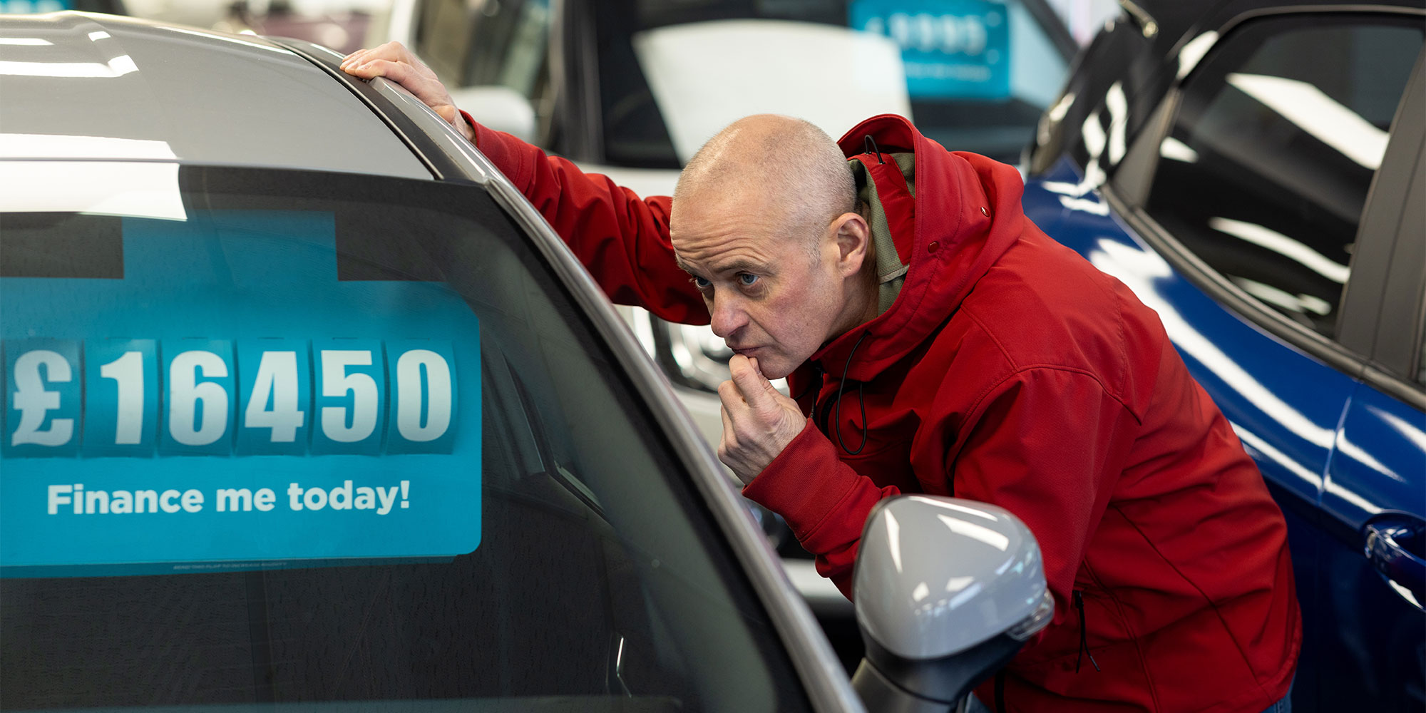 Man in a red coat looking at a car with a price tag in the window