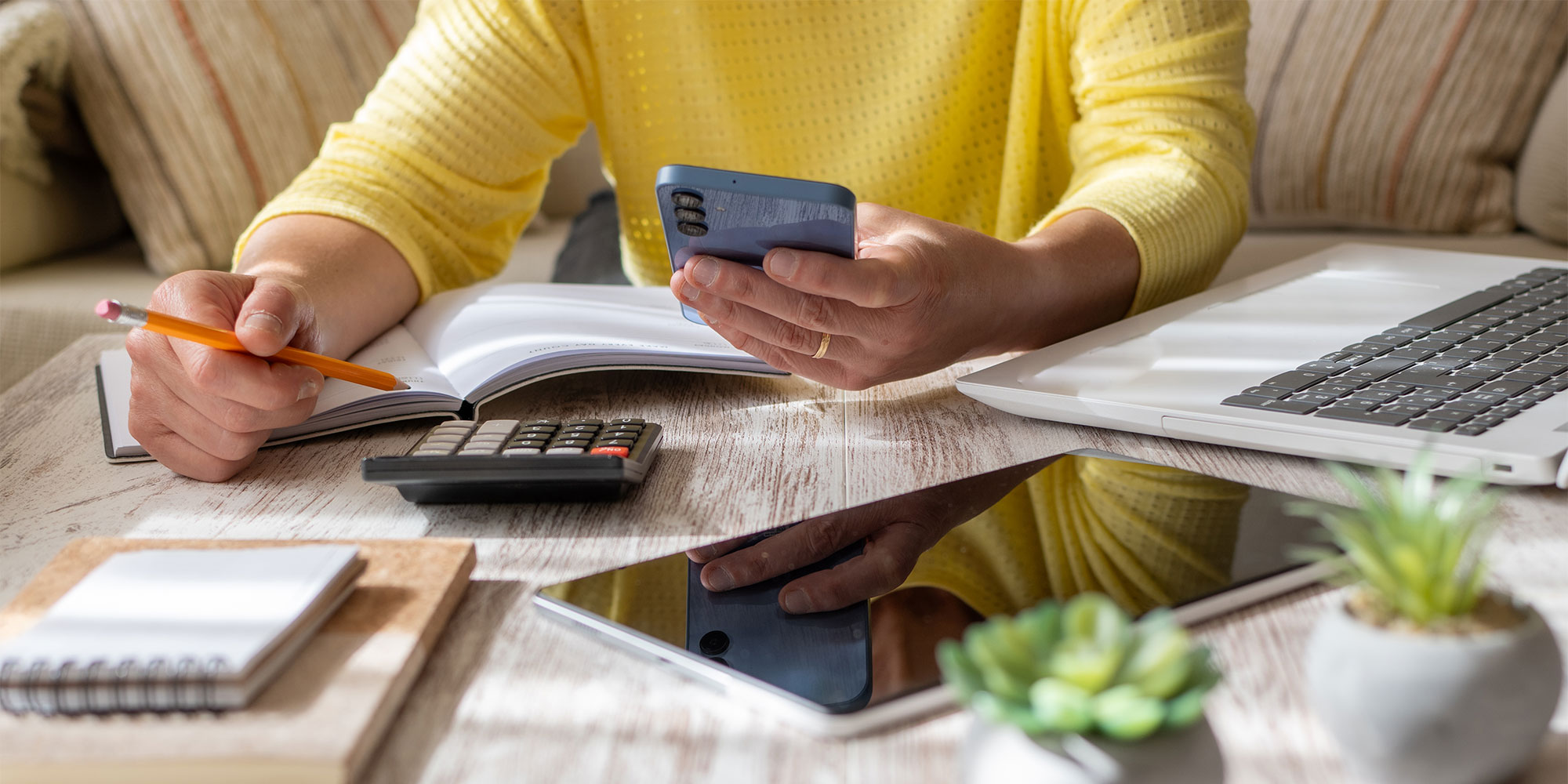 A person in a yellow sweater holds a phone and a pencil, surrounded by a notebook, calculator, tablet, and laptop.