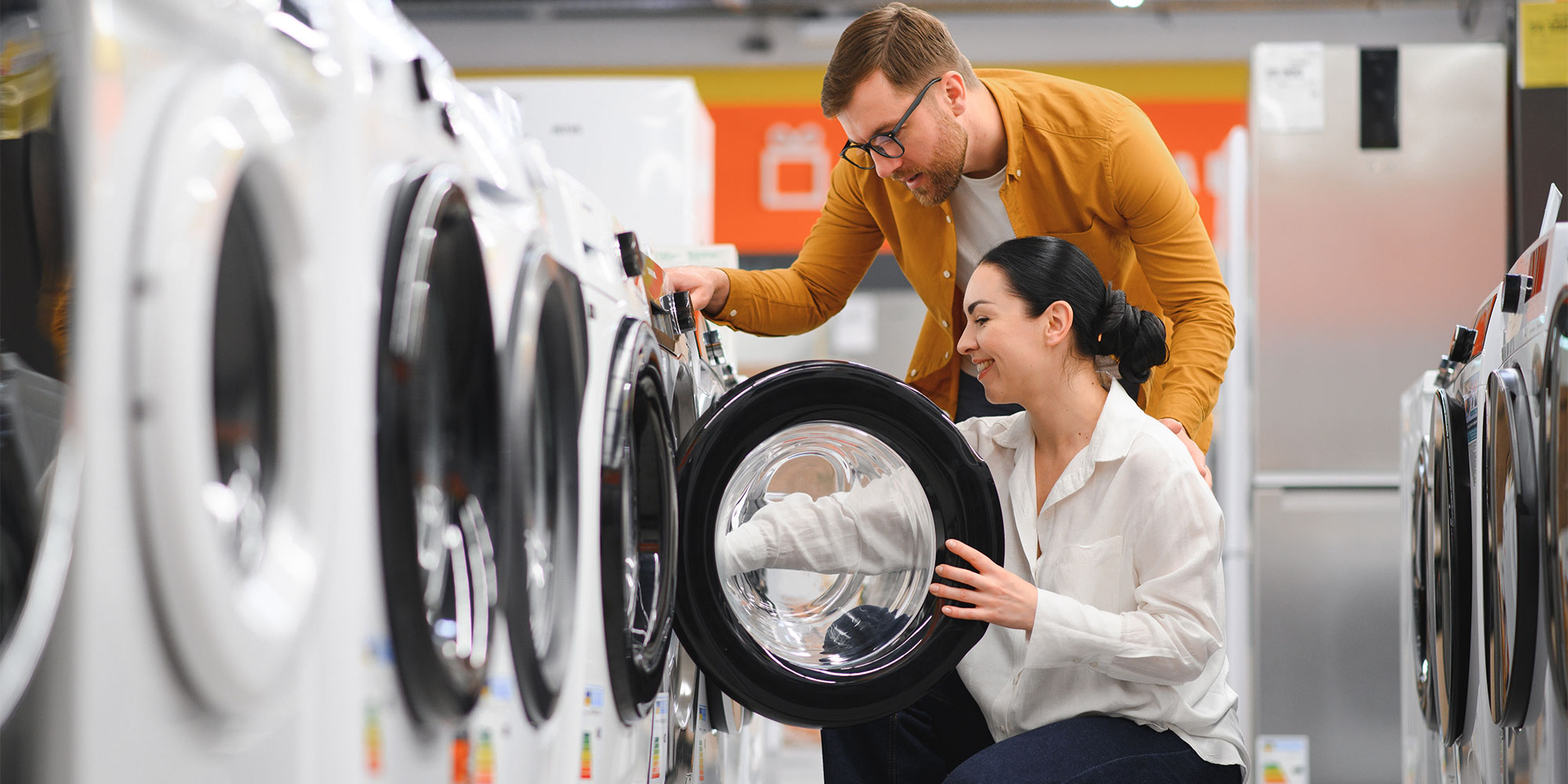 A man assists a woman inspecting a washing machine in an appliance store, surrounded by various models.