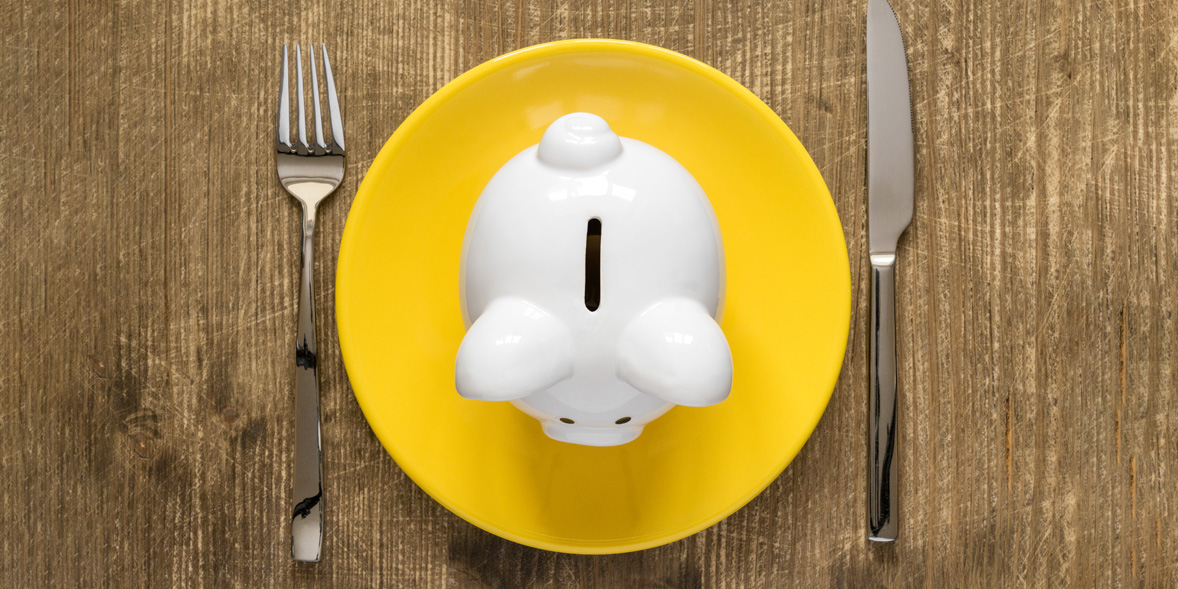White piggy bank on a bright yellow plate, surrounded by a fork and knife on a wooden table.