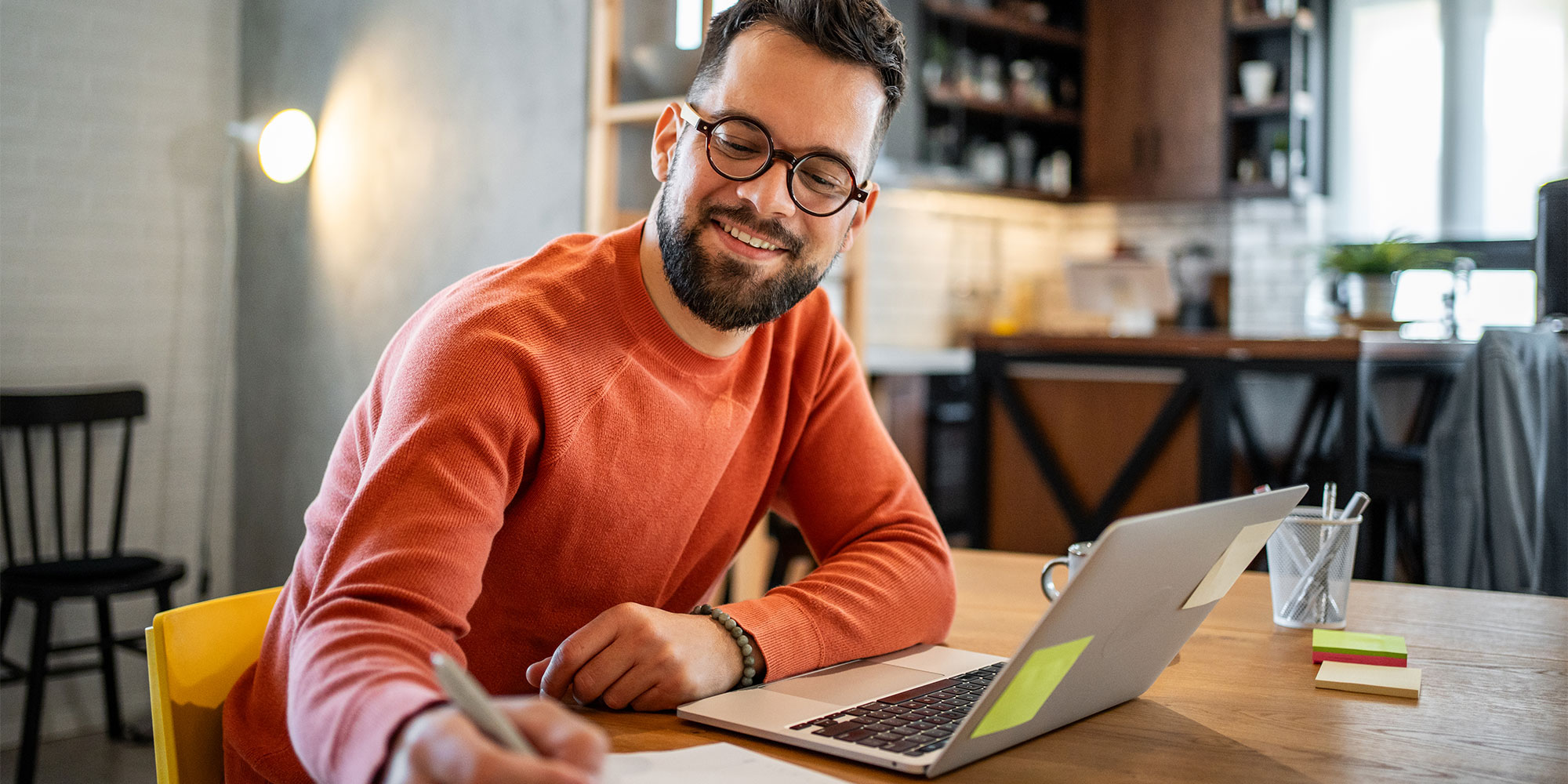 Man looks happy as he writes on paper alongside laptop