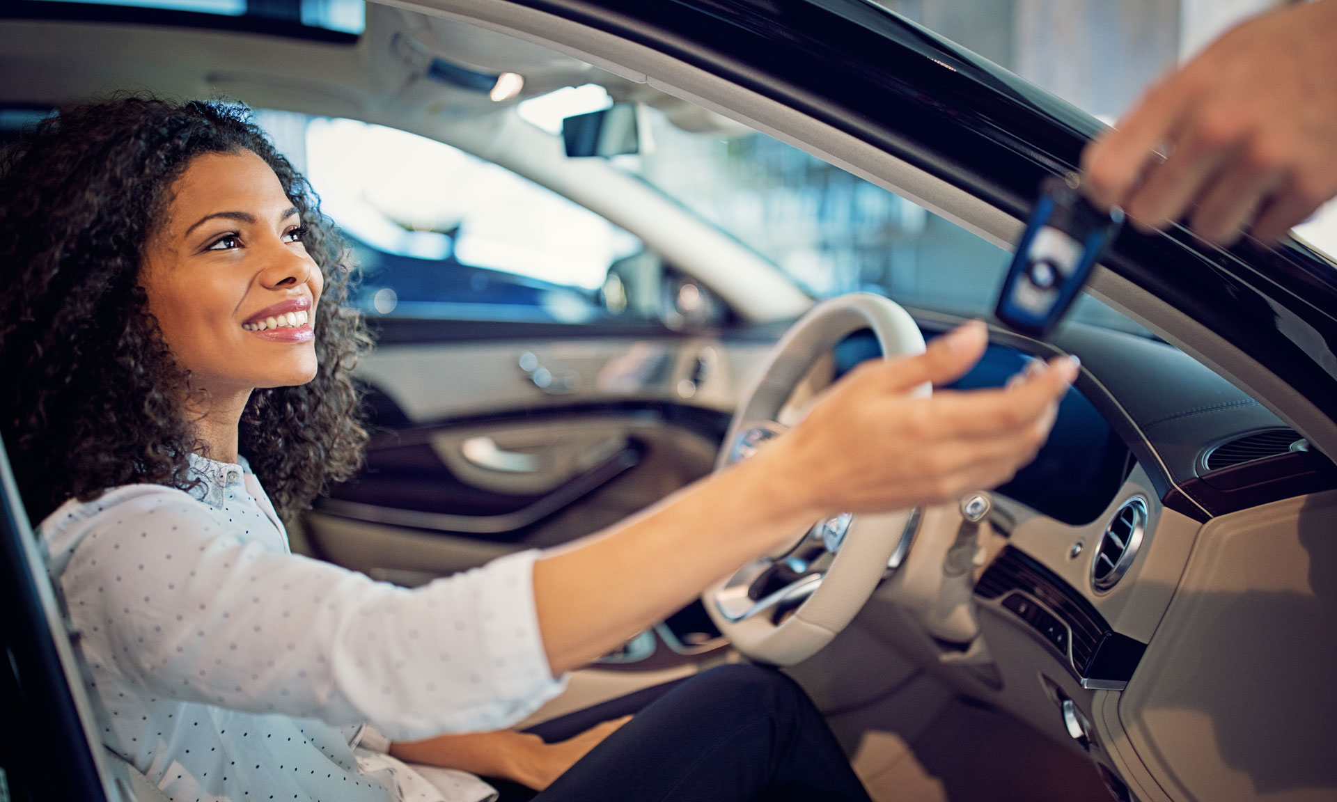 Young woman in a car, smiling and reaching out to receive a car key from a person outside the vehicle.