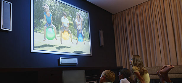Children watch a video on a large screen featuring kids playing on colorful inflatable balls outside.