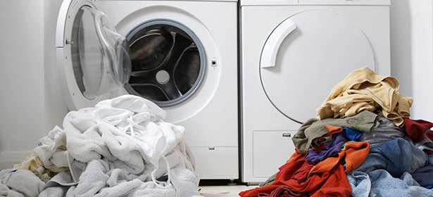 Front-loading washing machine with open door beside a dryer, stacked with piles of colorful and white laundry.