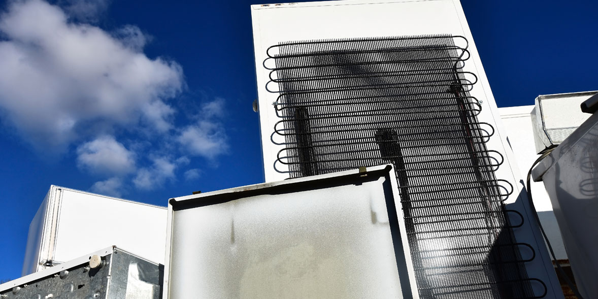 Bright blue sky contrasts with the white rear of a fridge with black coils on the surface.