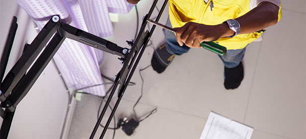 A person in a yellow shirt uses a screwdriver to adjust a black equipment stand, with tools and papers nearby.