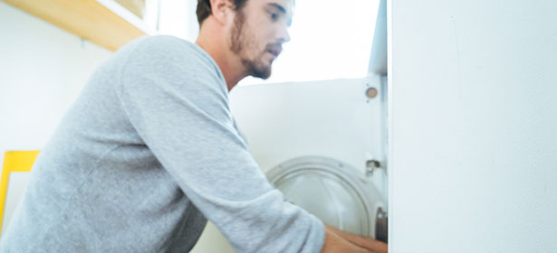 A man in a grey sweater is doing laundry, leaning over a washing machine in a well-lit laundry room.