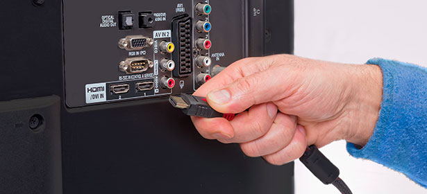 A hand is plugging an HDMI cable into the back of a television, near various audio and video input ports.