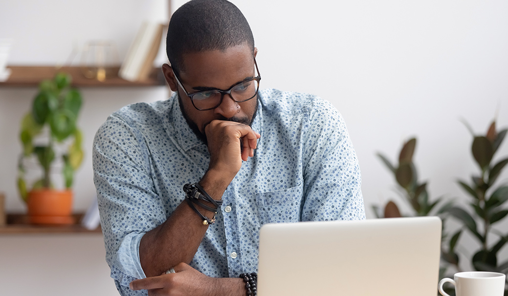 A man with glasses and a patterned blue shirt sits at a laptop, resting his chin on his hand, deep in thought.