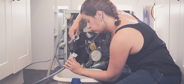 Woman in a tank top kneels by an open appliance, inspecting and handling pipes and components inside.