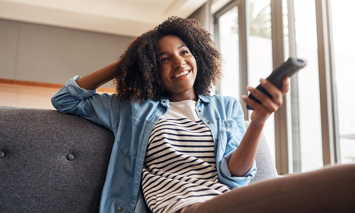 Smiling young woman with curly hair sits on a couch, holding a remote, enjoying a moment of relaxation at home.