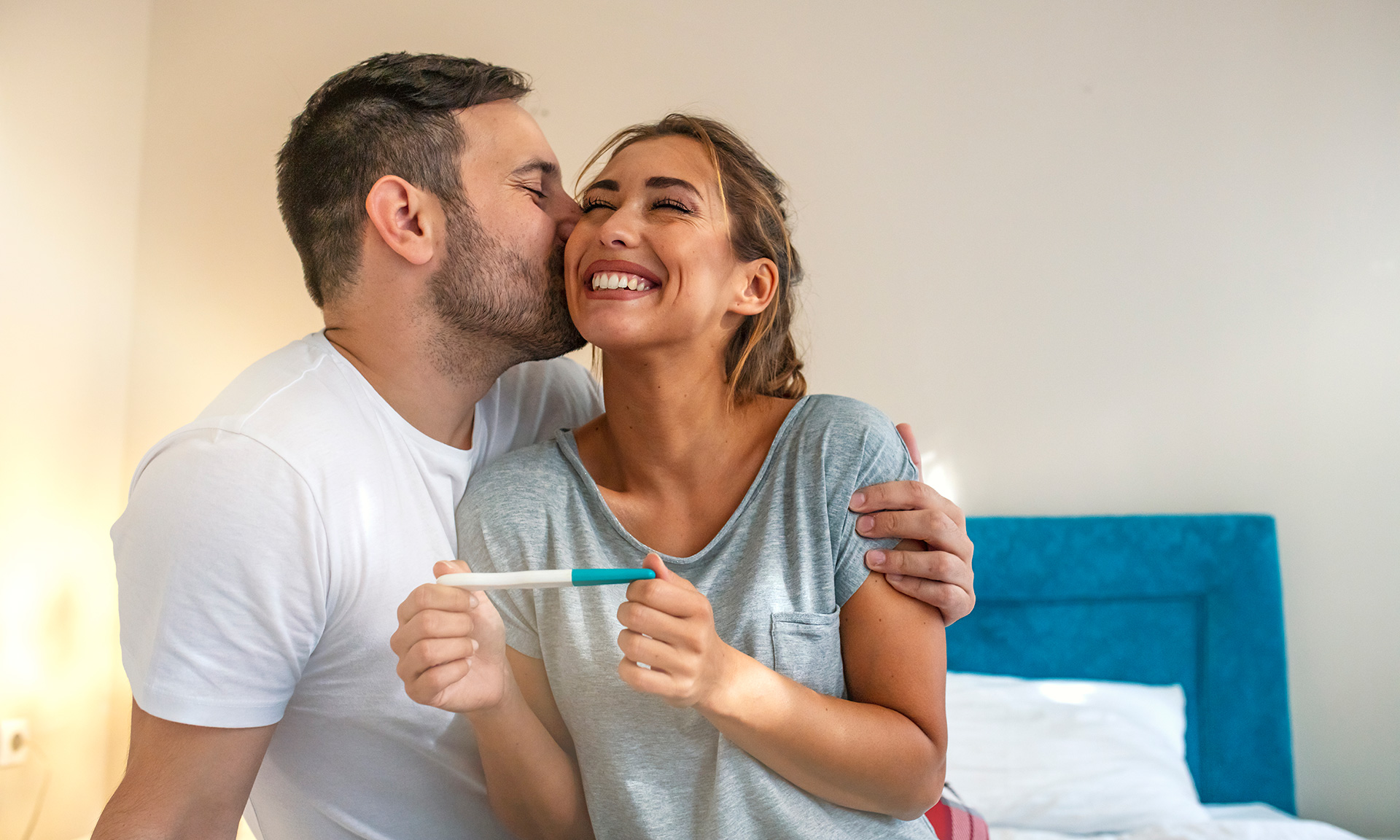 A couple embraces, smiling widely as the woman holds a pregnancy test, with a cozy bedroom setting in the background.