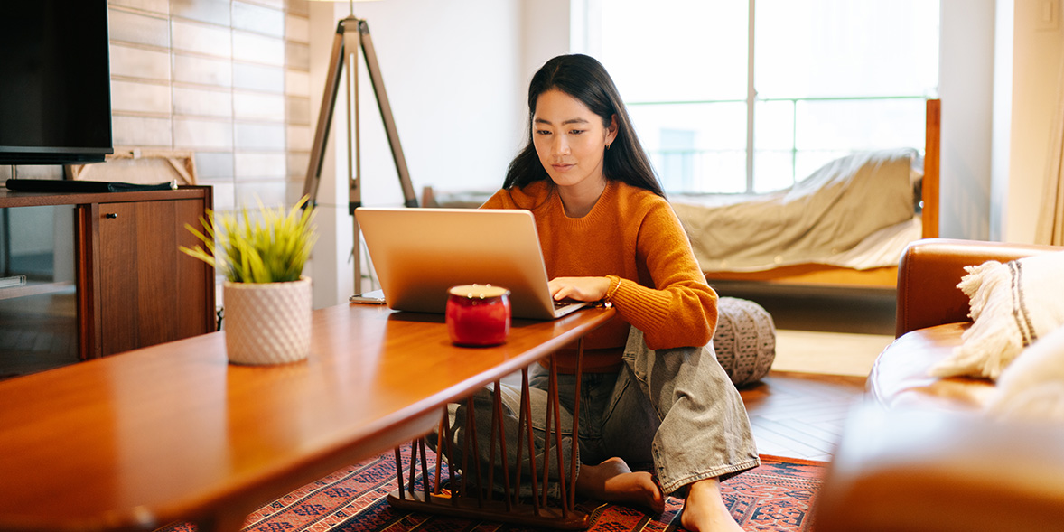 A woman with long black hair sits on the floor, using a laptop on a low table in a cozy, well-lit living room.