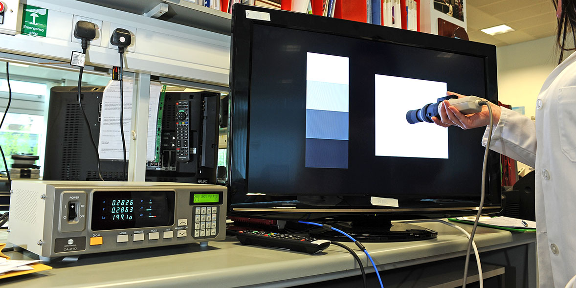 A monitor displays colour samples while a device is held to measure colour, with electronics equipment on the workspace.