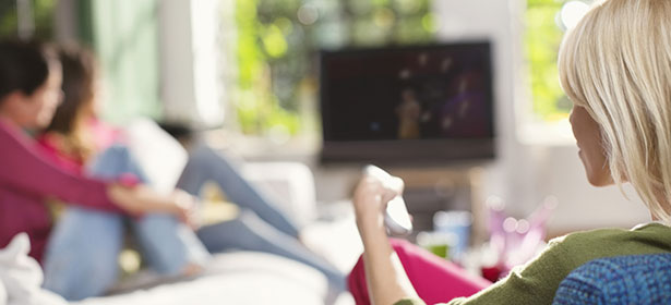 A cozy living room scene with two people relaxing on a sofa; one holding a remote, watching a TV in the background.