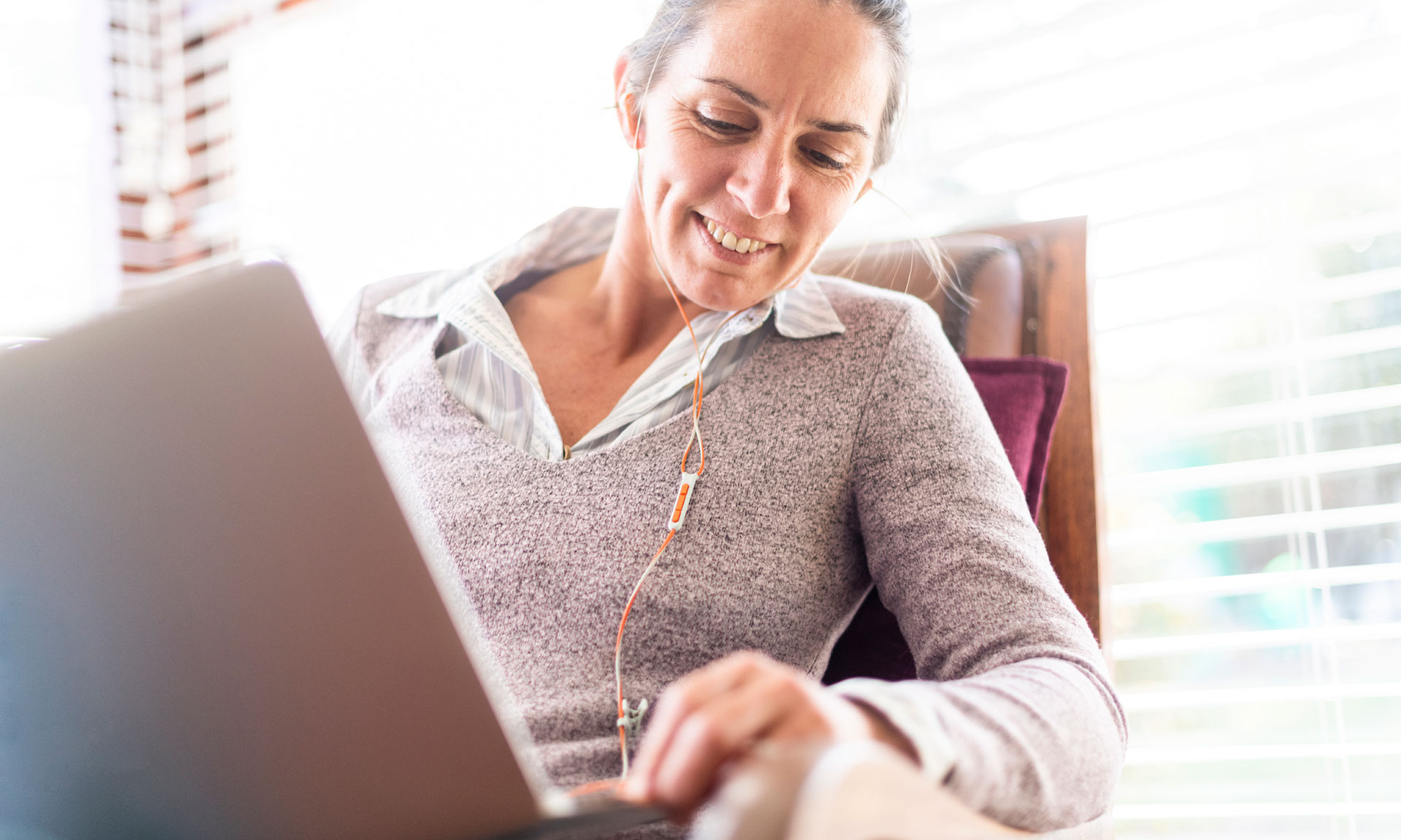 Woman with earphones smiles while working on a laptop, seated in a cozy chair near a window with natural light.