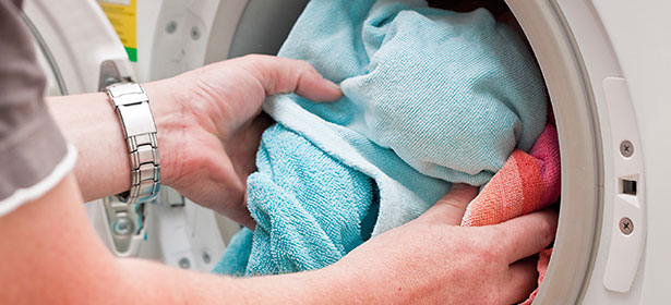 Hands reaching into a washing machine to place blue and pink towels inside for laundry.