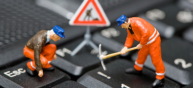 Two miniature construction workers are on a computer keyboard, one using a pickaxe, with a warning sign in the background.