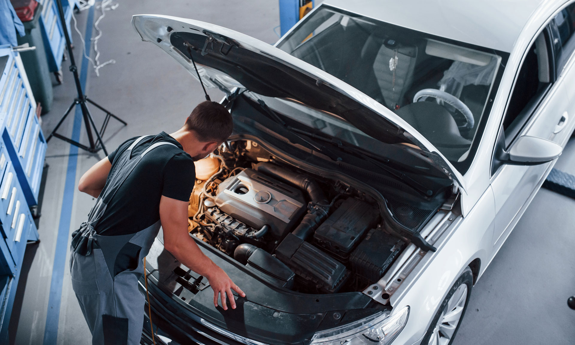 Mechanic inspecting the engine of a silver car with the hood raised, in a well-lit garage workshop.