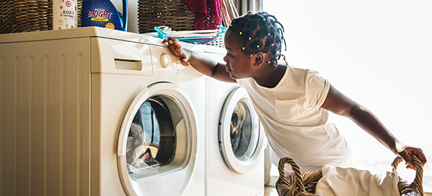 A young child with braided hair reaches towards a washing machine, curiously adjusting its controls in a bright laundry room.