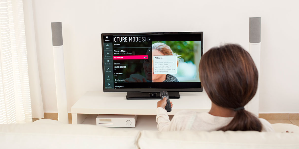 A person sits on a sofa, holding a remote, watching a TV displaying a menu screen with options and a video thumbnail.