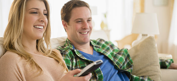 Smiling young couple relaxes on a couch, the woman holding a remote control while the man laughs at something on screen.