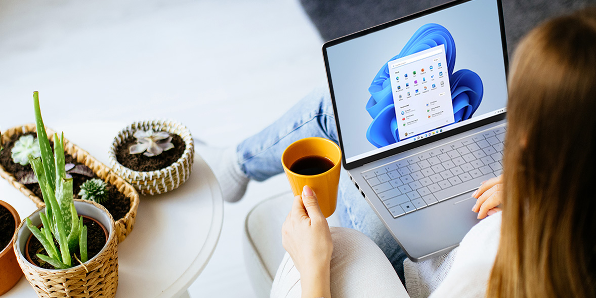 A person holds a yellow cup of coffee while using a laptop displaying Windows 11, surrounded by indoor plants.