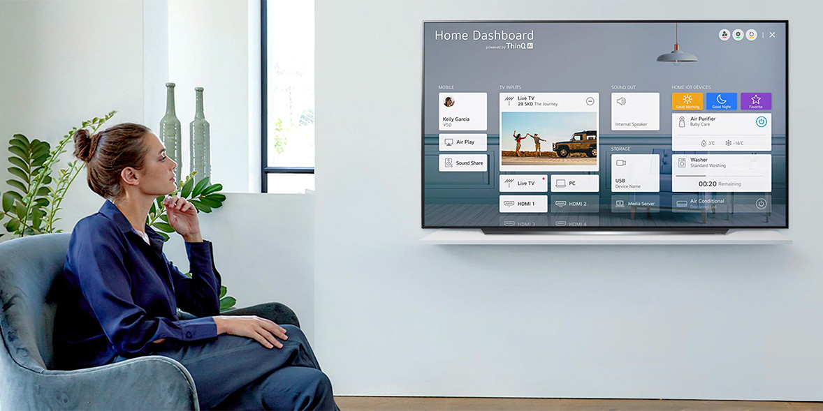 A woman in a navy blouse sits on a sofa, viewing a modern TV displaying a home dashboard interface with various apps.