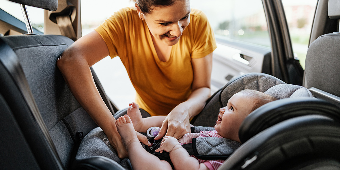 A smiling woman secures a laughing baby in a car seat inside a vehicle, with sunlight streaming through the window.