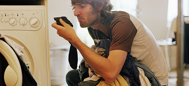 Man grimacing while smelling a wrinkled pile of laundry near a washing machine, surrounded by clothing in vibrant colors.