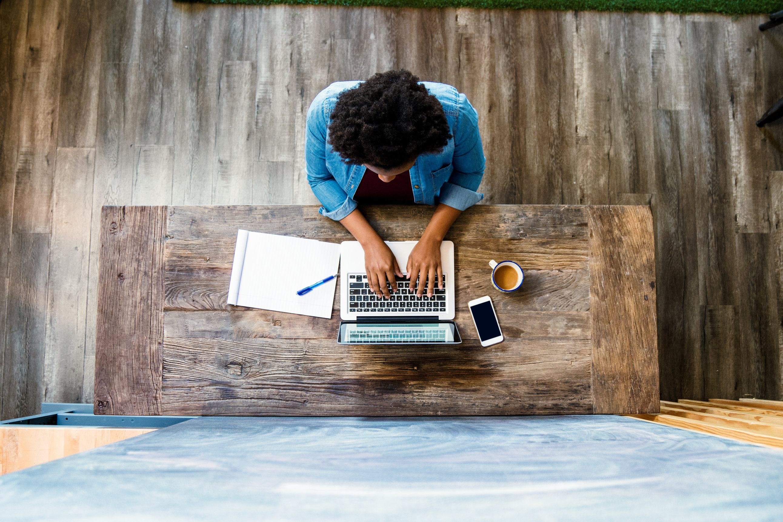 Person typing on a laptop at a wooden desk, with a notebook, pen, coffee cup, and smartphone nearby.