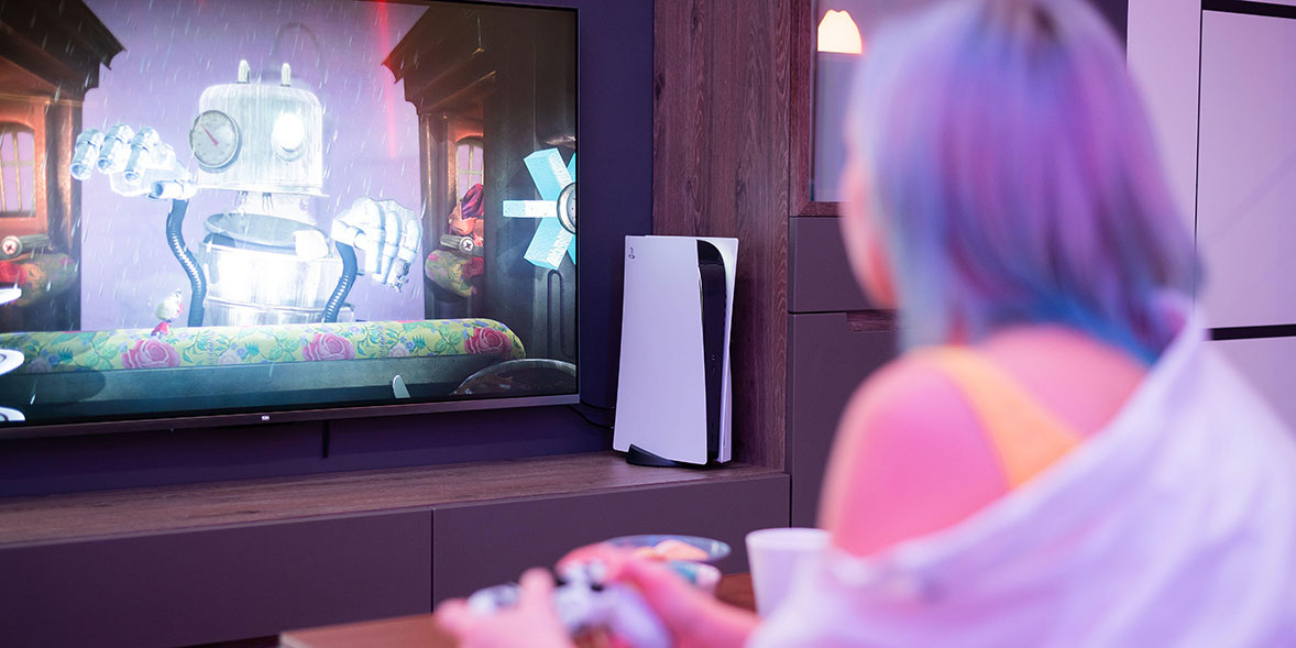 A person with pastel blue hair plays video games on a console while watching a brightly colored animated scene on a large TV.