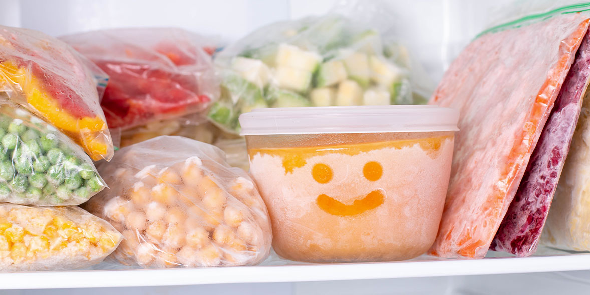 A container with a smiley face sits in a freezer surrounded by various bags of frozen vegetables and fruits.