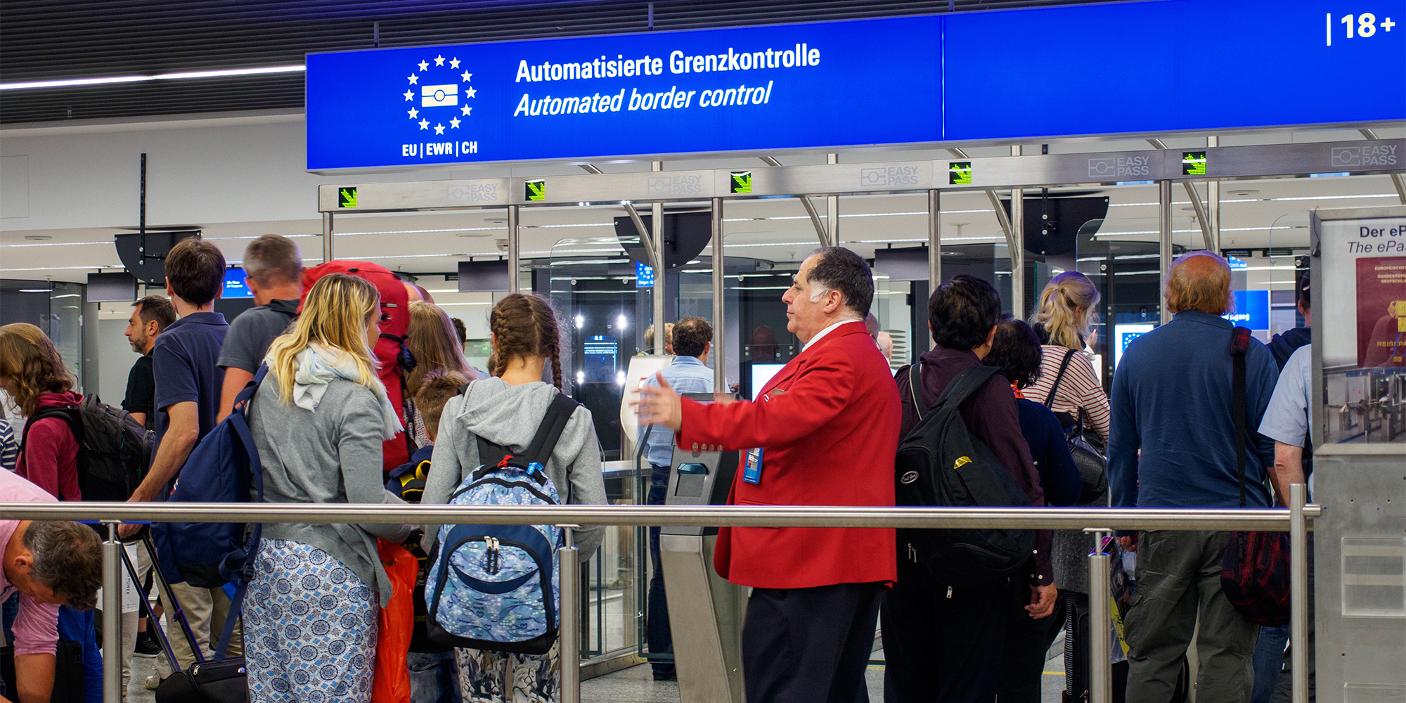 Travelers wait in line at a busy airport check-in area, with luggage carts and flight information displays in the background.